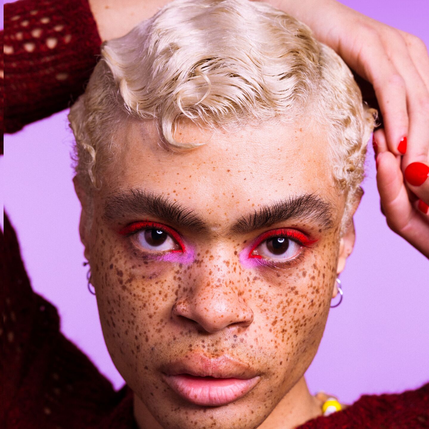 A person with short blond hair and freckles looks directly at the camera, wearing red and pink eyeshadow, with one hand touching their head. The background is lavender.