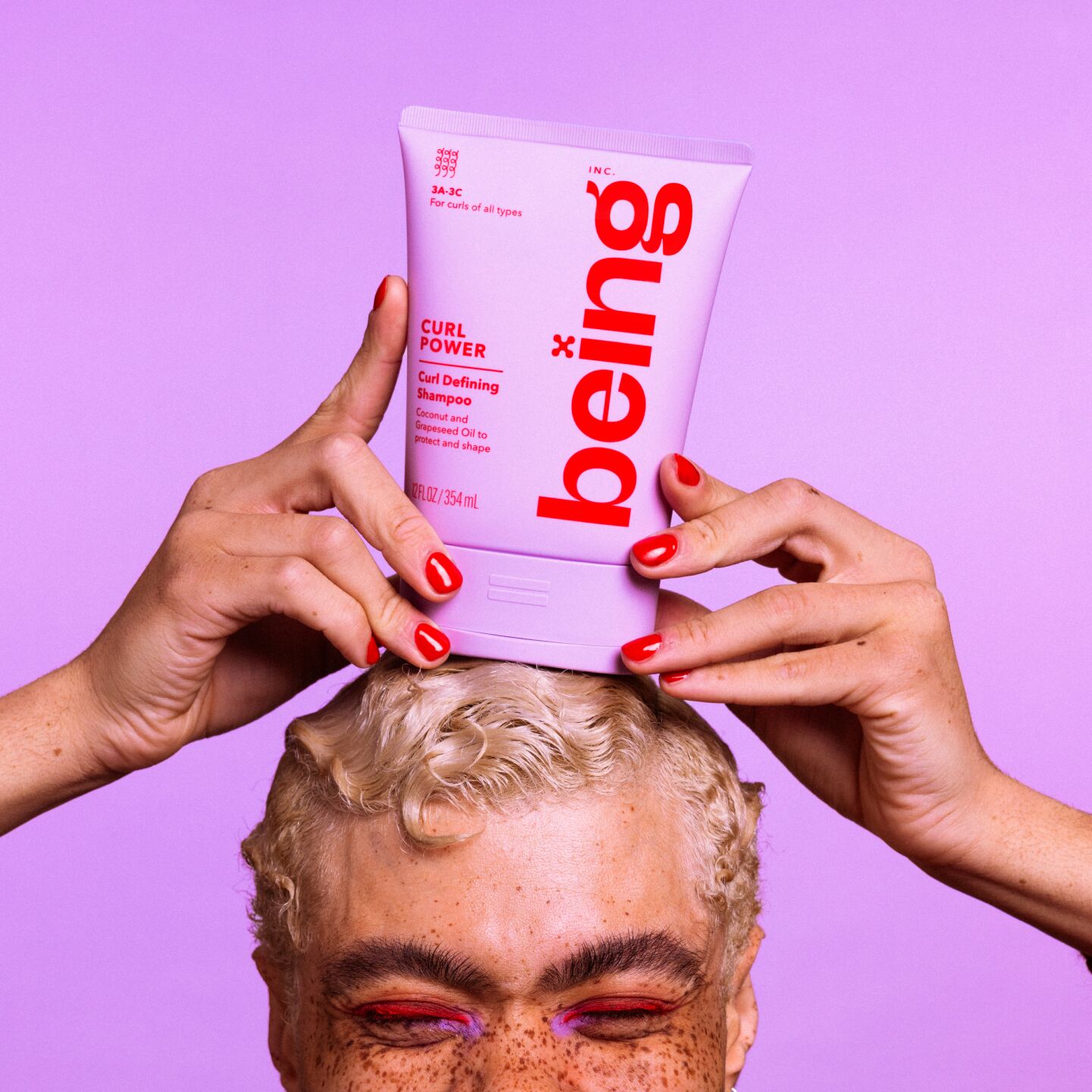 A person with short blond hair and freckles balances a a lavender bottle of 'being' Curl Power Curl Defining Shampoo on their head, against a lavender background.