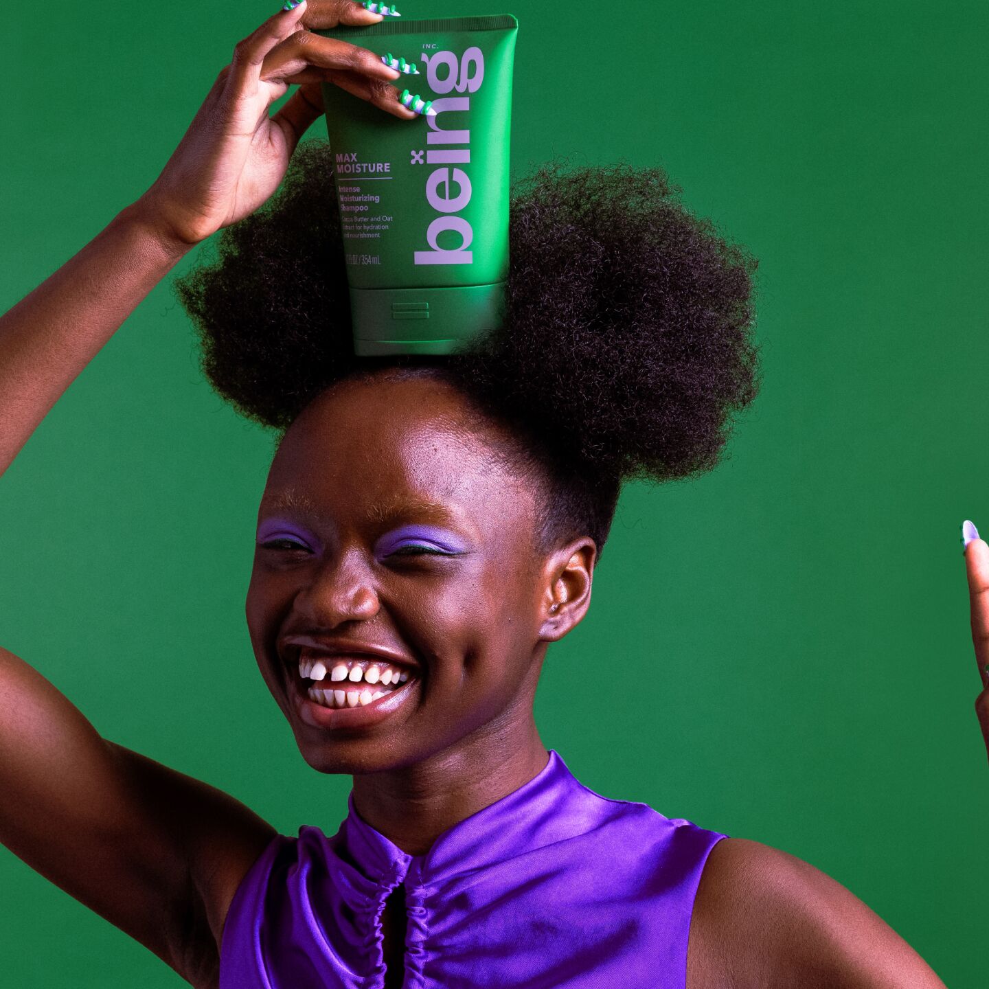 A smiling person with dark skin and two afro puffs holds a green bottle of 'being' Max Moisture Intense Moisturizing Shampoo on their head against a green background. They are wearing a purple top and have purple eyeshadow, with their nails painted green and white.