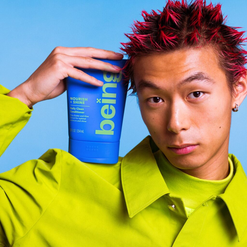 A person with spiky red hair holds a blue bottle of 'being' Nourish + Shine Daily Clean Conditioner against their head. They are wearing a green outfit, set against a blue background.