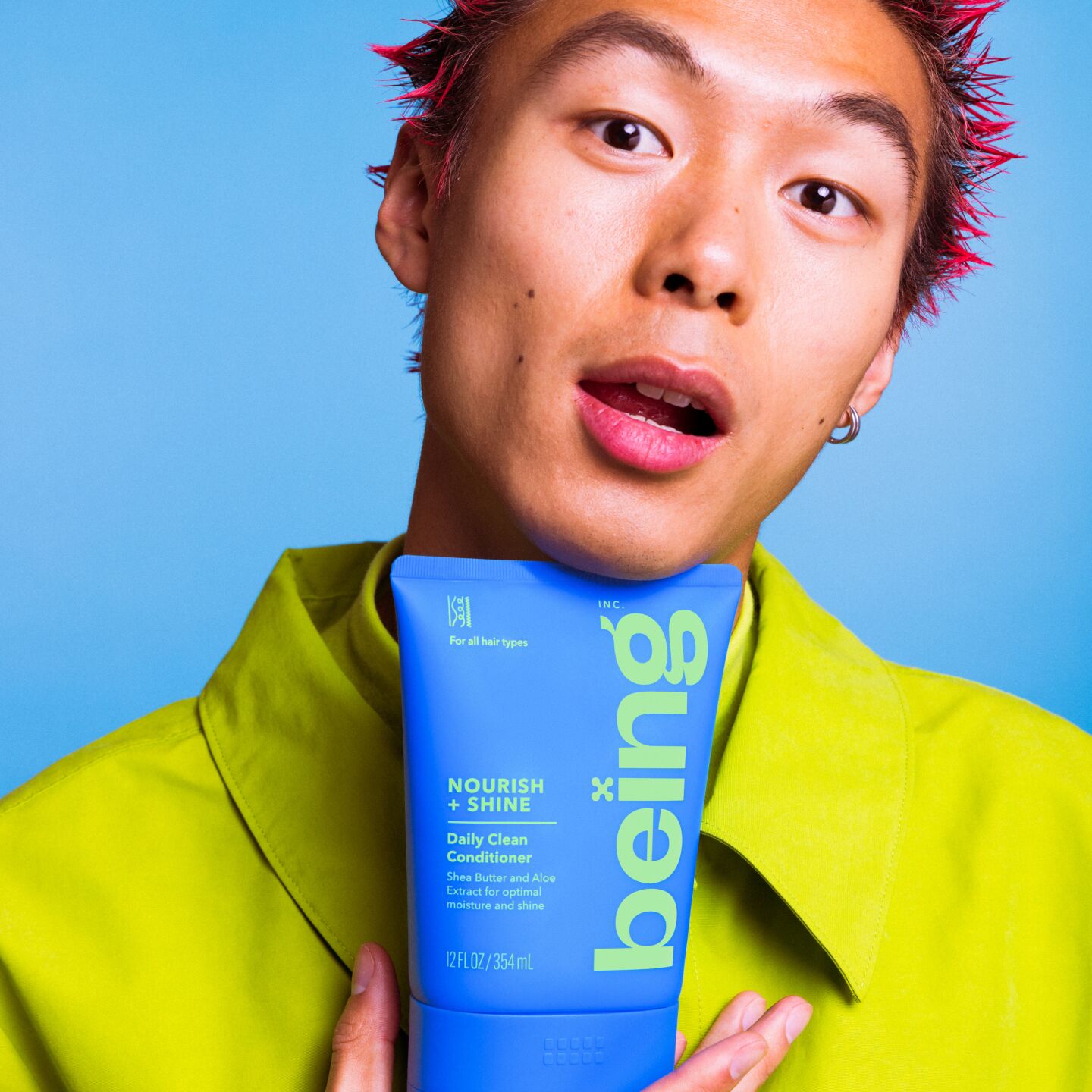 A person with spiky red hair holds a blue bottle of 'being' Nourish + Shine Daily Clean Conditioner against their chest. They are wearing a green outfit, set against a blue background.