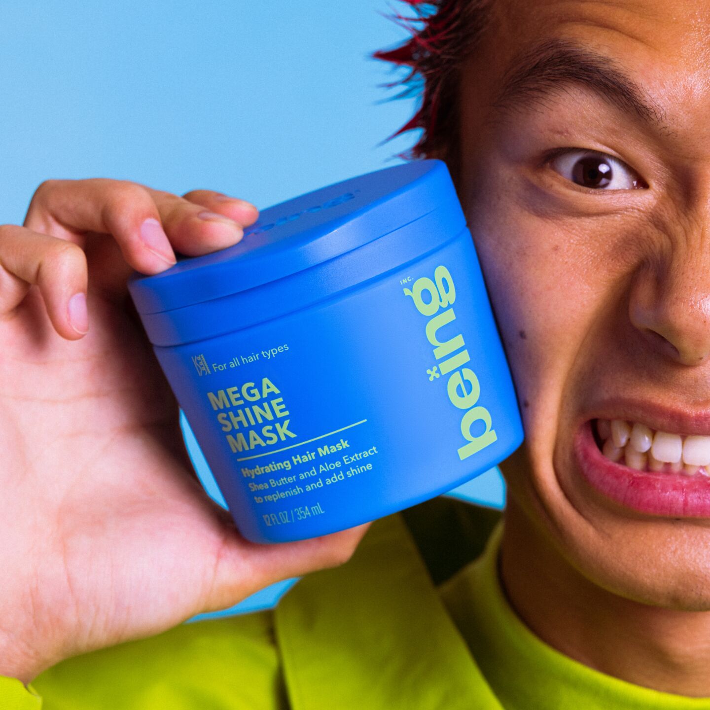 A person with spiky red hair makes a scrunched-up facial expression while holding a blue jar of 'being' Mega Shine Mask Hydrating Hair Mask against their cheek. They are wearing a green outfit, set against a blue background.