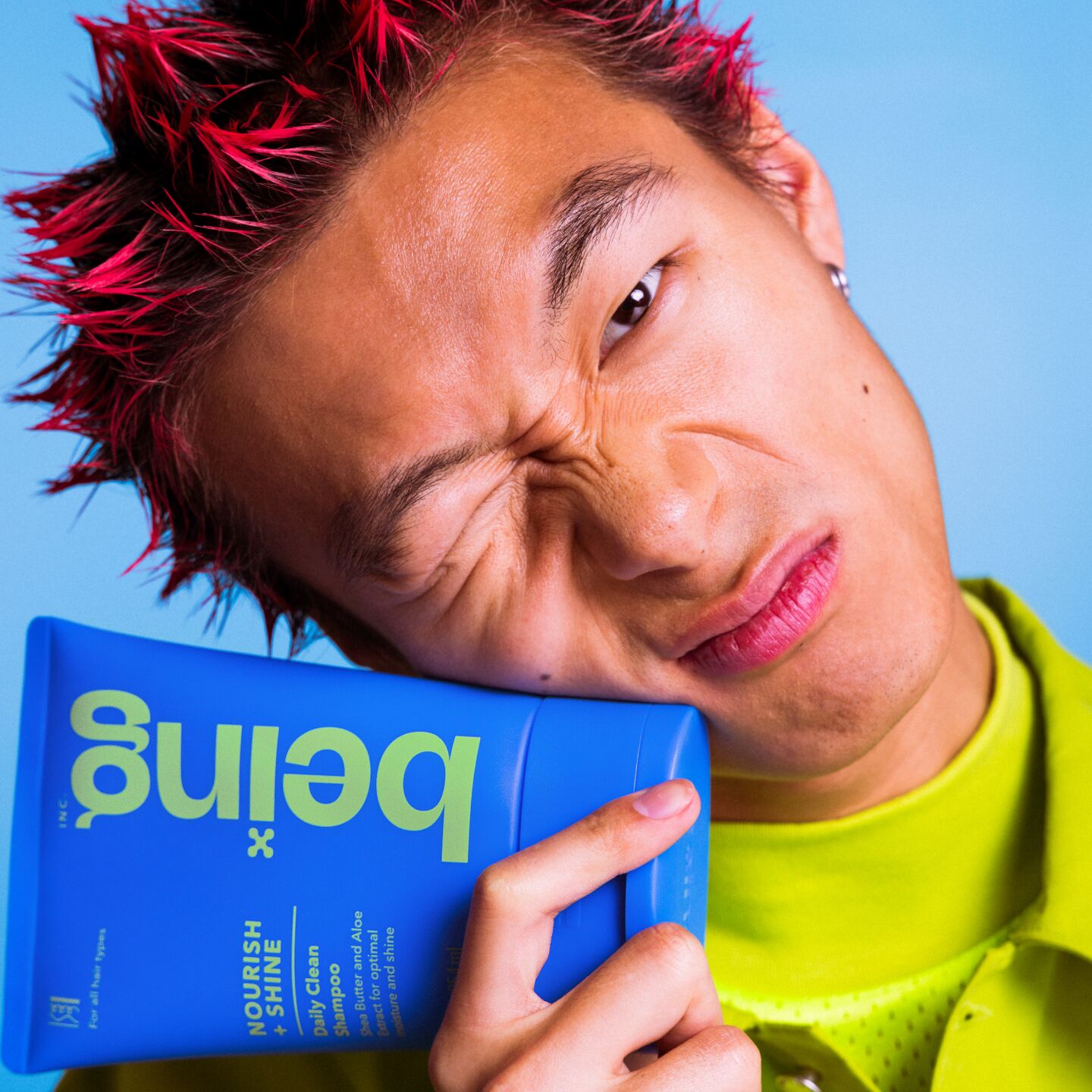 A person with spiky red hair makes a scrunched-up facial expression while holding a blue bottle of 'being' Nourish + Shine Daily Clean Shampoo against their cheek. They are wearing a green outfit, set against a blue background.