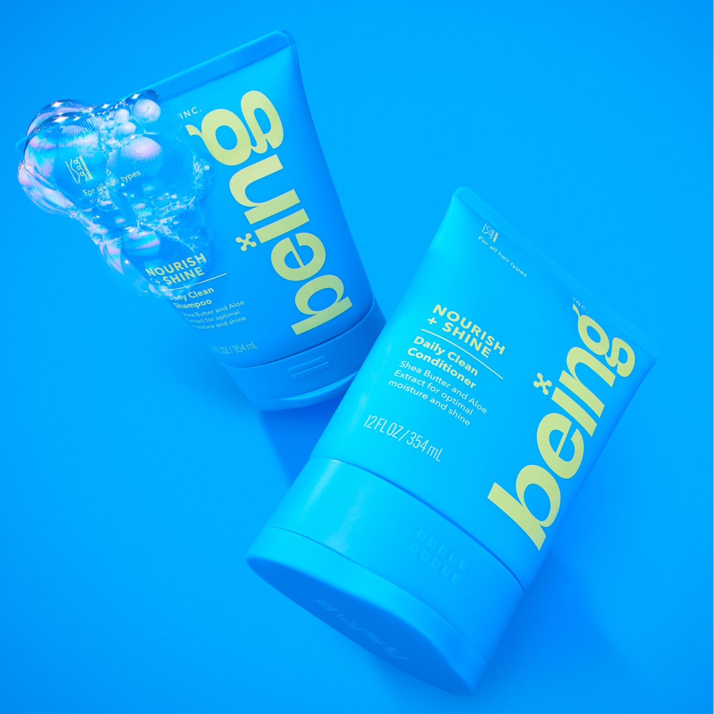 Two blue bottles of 'being' Nourish + Shine Daily Clean Shampoo and Conditioner, with bubbles on the shampoo bottle, set against a blue background.