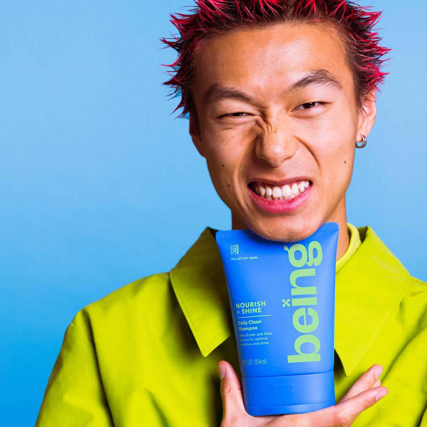 A person with spiky red hair makes a scrunched-up facial expression while holding a blue bottle of 'being' Nourish + Shine Daily Clean Shampoo against their chest. They are wearing a green outfit, set against a blue background.