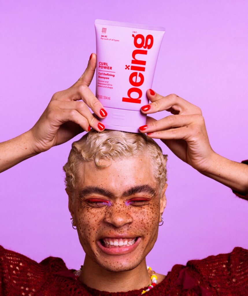 A person with short blond hair and freckles balances a a lavender bottle of 'being' Curl Power Curl Defining Shampoo on their head, against a lavender background.