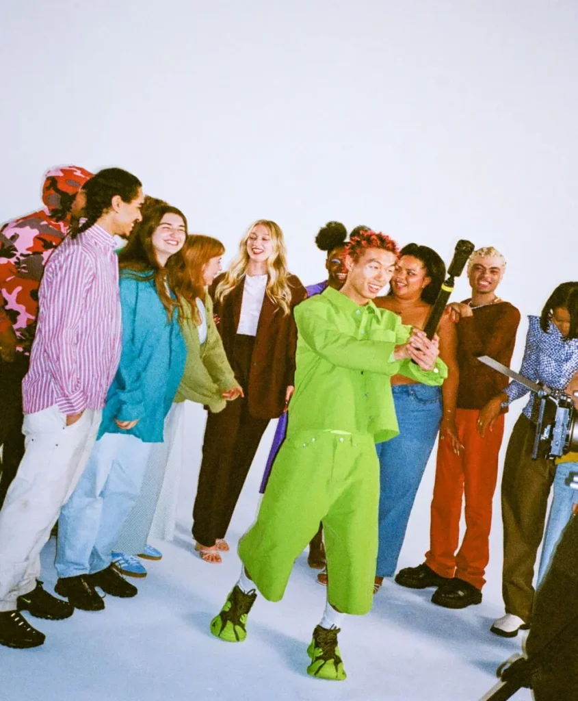 A group of people standing together in a studio setting, with one person in a bright green outfit holding a camera or similar object, and others smiling and interacting around them.